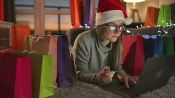 Happy woman with glasses wearing a santa claus hat is lying on the carpet and makes an online purchase using a credit card video