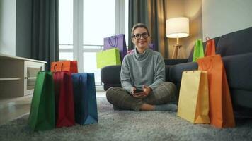 Porträt von ein glücklich Frau Sitzung auf ein Teppich im ein gemütlich Zimmer unter Einkaufen Taschen video
