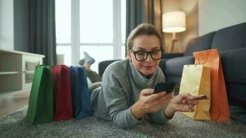 Happy woman is lying on the floor and makes an online purchase using a credit card and smartphone. Shopping bags around video