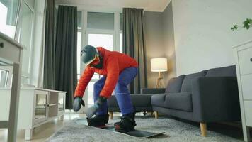 pret video. Mens gekleed net zo een snowboarder ritten een snowboard Aan een tapijt in een knus kamer. aan het wachten voor een besneeuwd winter video