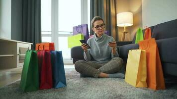 feliz mulher com óculos é sentado em a tapete e faz a conectados compra usando uma crédito cartão e Smartphone. compras bolsas em volta. video
