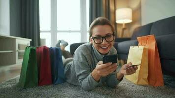 glücklich Frau ist Lügen auf das Fußboden und macht ein online Kauf mit ein Anerkennung Karte und Smartphone. Einkaufen Taschen um video