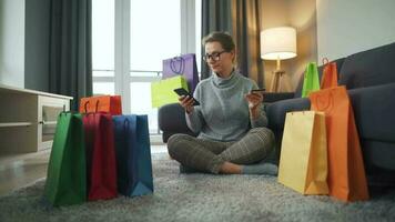 Happy woman with glasses is sitting on the carpet and makes an online purchase using a credit card and smartphone. Shopping bags around. video