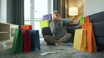 Happy woman is sitting on a carpet in a cozy room among shopping bags and making money rain from US dollar bills video