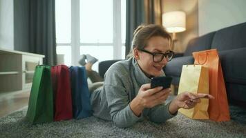 glücklich Frau ist Lügen auf das Fußboden und macht ein online Kauf mit ein Anerkennung Karte und Smartphone. Einkaufen Taschen um video