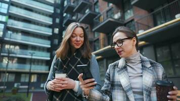 Two happy women walking with takeaway coffee and talking with interest among themselves in the business district. Slow video