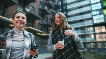 Two happy women walking with takeaway coffee and talking with interest among themselves in the business district. Slow video