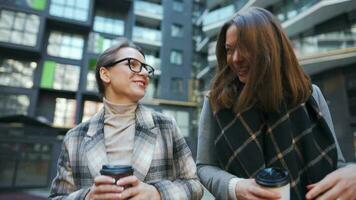 Two happy women walking with takeaway coffee and talking with interest among themselves in the business district. Slow motion video