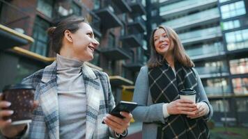 Two happy women walking with takeaway coffee and talking with interest among themselves in the business district. Slow video