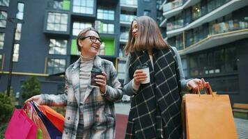 zwei glücklich Frauen gehen mit Einkaufen Taschen und wegbringen Kaffee nach ein erfolgreich Einkaufen und sich unterhalten mit Interesse unter sich video
