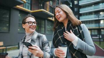 Two happy women walking with takeaway coffee and talking with interest among themselves in the business district. Slow video