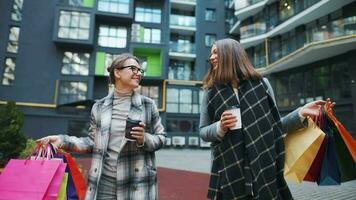 Two happy women walk with shopping bags and takeaway coffee after a successful shopping and talk with interest among themselves video