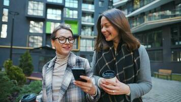 Two happy women walking with takeaway coffee and talking with interest among themselves in the business district. Slow video