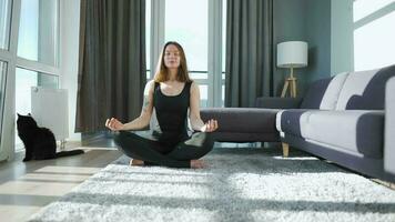 Young caucasian woman in black jumpsuit sitting in lotus position and meditating, yoga at home. Black cat walks around video