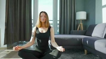 Jeune caucasien femme dans noir combinaison séance dans lotus position et méditer, yoga à maison. lent mouvement video