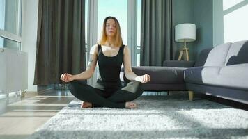 Young caucasian woman in black jumpsuit sitting in lotus position and meditating, yoga at home. Slow motion video