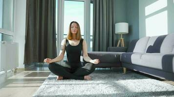 Young caucasian woman in black jumpsuit sitting in lotus position and meditating, yoga at home. Slow motion video