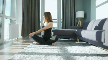 jong Kaukasisch vrouw in zwart jumpsuit zittend in lotus positie en mediteren, yoga Bij huis. langzaam beweging video
