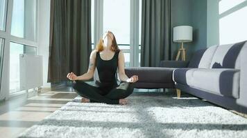 Young caucasian woman in black jumpsuit sitting in lotus position and meditating, yoga at home. Slow motion video