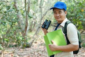 Asian man explorer wears blue cap, holds binocular in forest to survey botanical plants and creatures wildlife. Concept, nature exploration. Ecology and Environment. photo