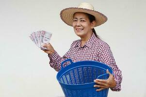 contento mayor mujer granjero usa sombrero, tartán camisa, sostiene tailandés billetes dinero y azul cesta. concepto, granjero contento a obtener ganancia, ingresos desde haciendo agricultura. obtener secundario dinero. foto