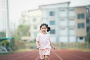 bebé asiático niña correr trotar a corriendo pista, correr campo a estadio. linda hembra ejercicio a puesta de sol contento bebé niña sonriendo a puesta de sol. linda bebé a corriendo pista. foto