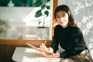 mujer trabajadora asiática con camisa negra y diario de escritura en un pequeño cuaderno en la mesa en el café interior. notas de mujer y tomando café en la cafetería. trabajar desde cualquier lugar concepto. foto