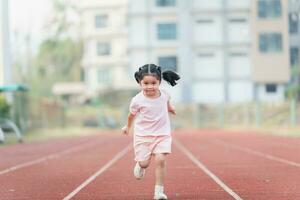 bebé asiático niña correr trotar a corriendo pista, correr campo a estadio. linda hembra ejercicio a puesta de sol contento bebé niña sonriendo a puesta de sol. linda bebé a corriendo pista. foto