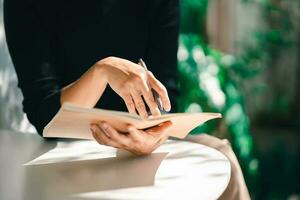 working woman wearing black shirt and writing journal on small notebook on the table at indoor cafe. Woman notes and drinking coffee at cafe. Working from anywhere concept. photo