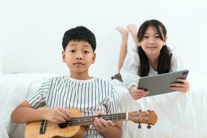 Brother holding an ukulele, sister holding a tablet looking at the camera in the bedroom photo