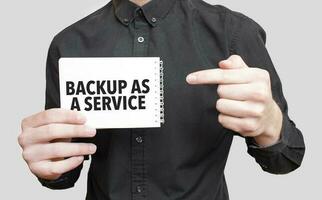 Businessman holding a white notepad with text BACKUP AS A SERVICE, business concept photo