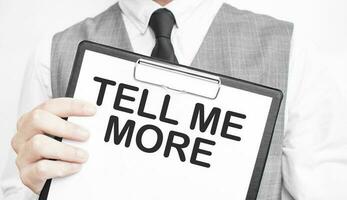 TELL ME MORE inscription on a notebook in the hands of a businessman on a grey background, a man points with a finger to the text photo