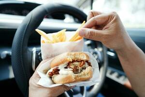 asiático mujer conductor sostener y comer hamburguesa y francés papas fritas en auto, peligroso y riesgo un accidente. foto