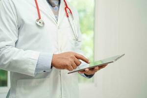 Asian man doctor using tablet computer to reading patient report, check up and search for solve treatment health medical online in hospital. photo