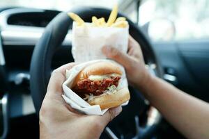 asiático mujer conductor sostener y comer hamburguesa y francés papas fritas en auto, peligroso y riesgo un accidente. foto