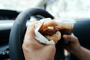 asiático mujer conductor sostener y comer hamburguesa en auto, peligroso y riesgo un accidente. foto