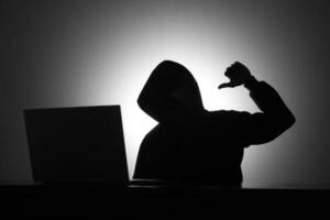 Mysterious man wearing black hoodie using laptop while mocking someone by making thumb down gesture in the dark room. Hacker and cyber security concept. photo