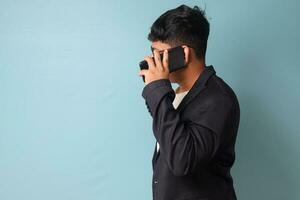 retrato de joven asiático negocio hombre en casual traje frente a lado mientras hablando con alguien en teléfono. aislado imagen en azul antecedentes foto