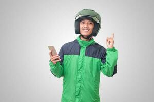 Portrait of Asian online taxi driver wearing green jacket and helmet holding mobile phone and pointing to empty space with finger. Isolated image on white background photo