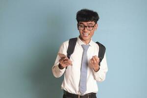 indonesio mayor alto colegio estudiante vistiendo blanco camisa uniforme con gris Corbata levantamiento su puño, celebrando éxito mientras participación un móvil teléfono. aislado imagen en azul antecedentes foto