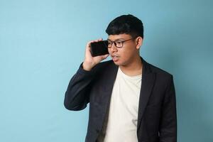 Portrait of young Asian business man in casual suit talking with someone on the phone. Isolated image on blue background photo