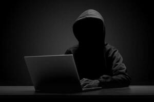 misterioso hombre vistiendo negro capucha utilizando ordenador portátil en el oscuro habitación. hacker y ciber seguridad concepto. foto