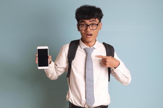 Indonesian senior high school student wearing white shirt uniform with gray tie showing and presenting blank screen mobile phone while pointing on his smartphone. Isolated image on blue background photo