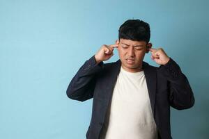 Portrait of young Asian business man in casual suit overwhelmed with noises and while closing ears with both hands. Isolated image on blue background photo