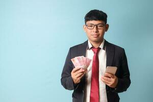 Portrait of young Asian business man in casual suit holding phone and thousand rupiahs looking at camera while smiling. Isolated image on blue background photo