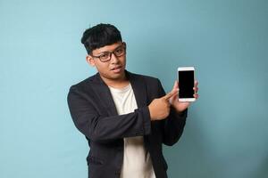 Portrait of young Asian business man in casual suit pointing at phone with confused expressions. Isolated image on blue background photo