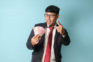 Portrait of young Asian business man in casual suit pointing hand at camera while holding thousand rupiahs with persuasive expressions. Isolated image on blue background photo