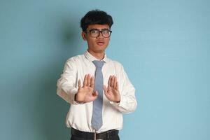 Indonesian senior high school student wearing white shirt uniform with gray tie forming a hand gesture to avoid something. Isolated image on blue background photo