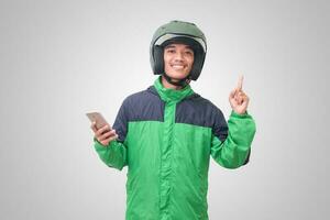 Portrait of Asian online taxi driver wearing green jacket and helmet holding mobile phone and pointing to empty space with finger. Isolated image on white background photo