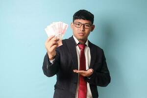 Portrait of young Asian business man in casual suit showing thousand rupiahs and making hand gestures with serious expressions. Isolated image on blue background photo